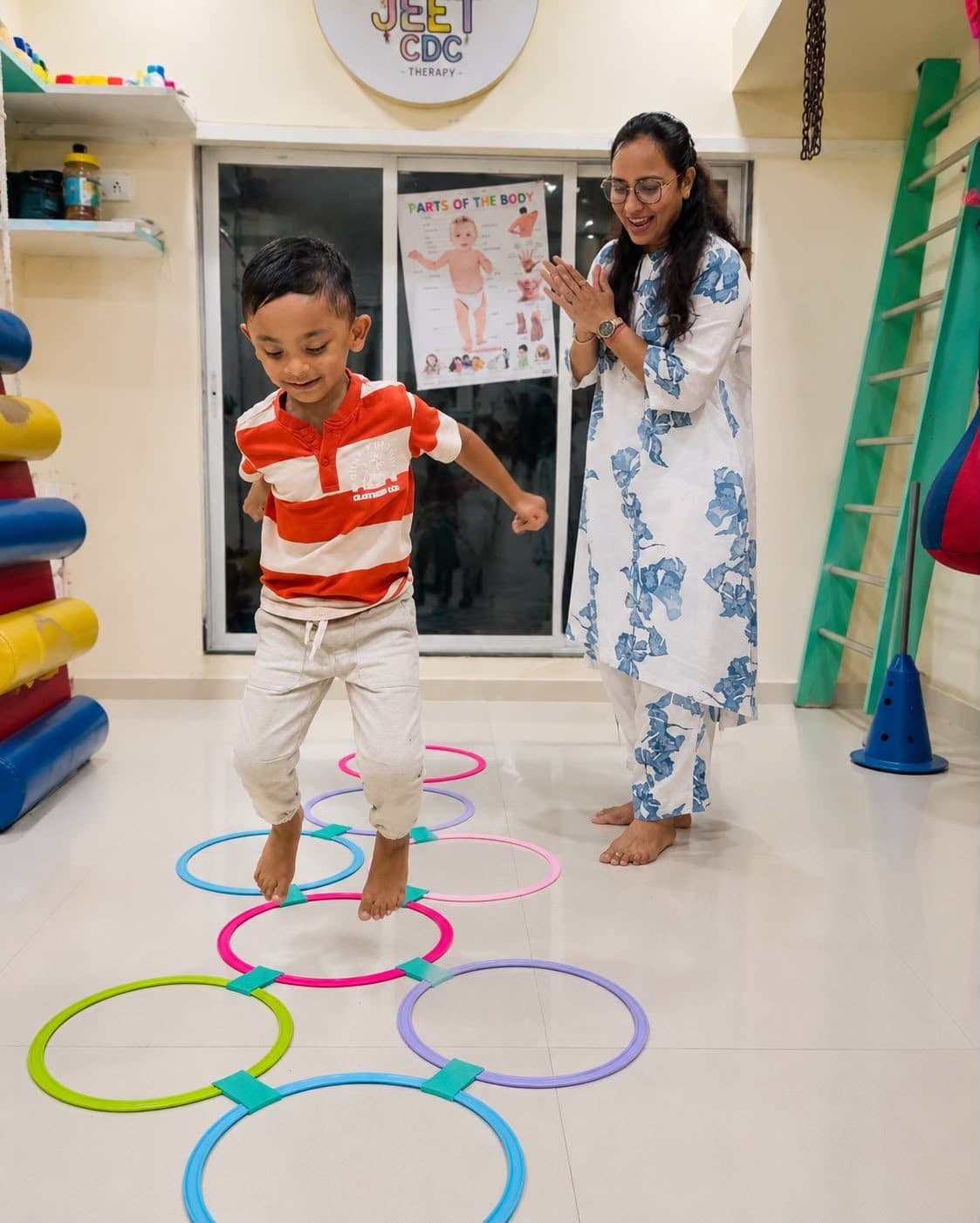 A boy jumping through colourful floor rings as the therapist cheers him on